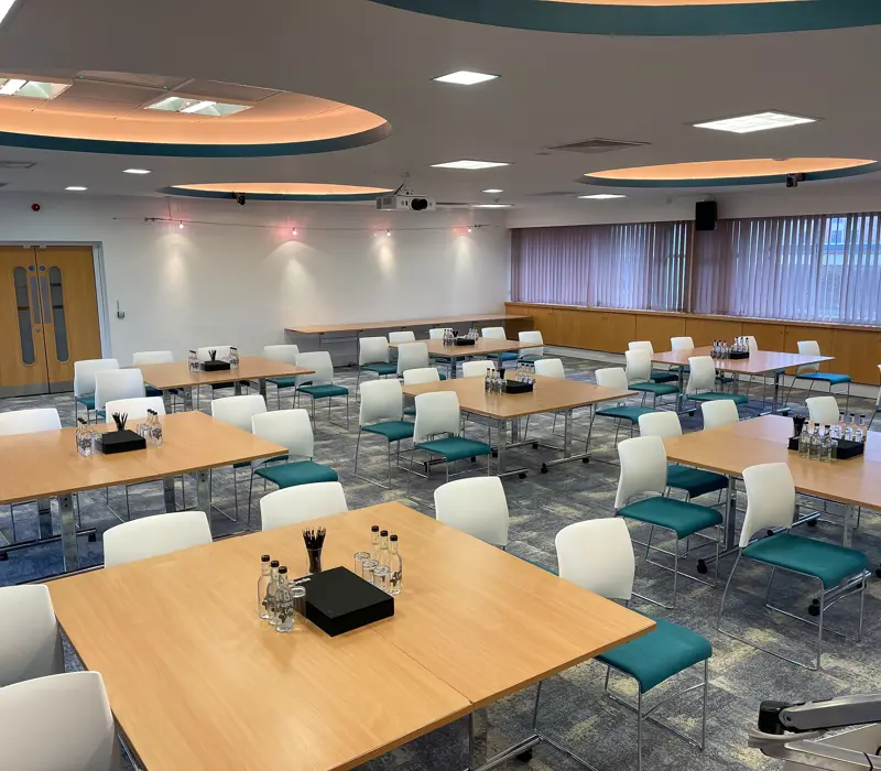 A modern conference room with multiple tables, teal and white chairs, set for a meeting with water bottles and stationery.
