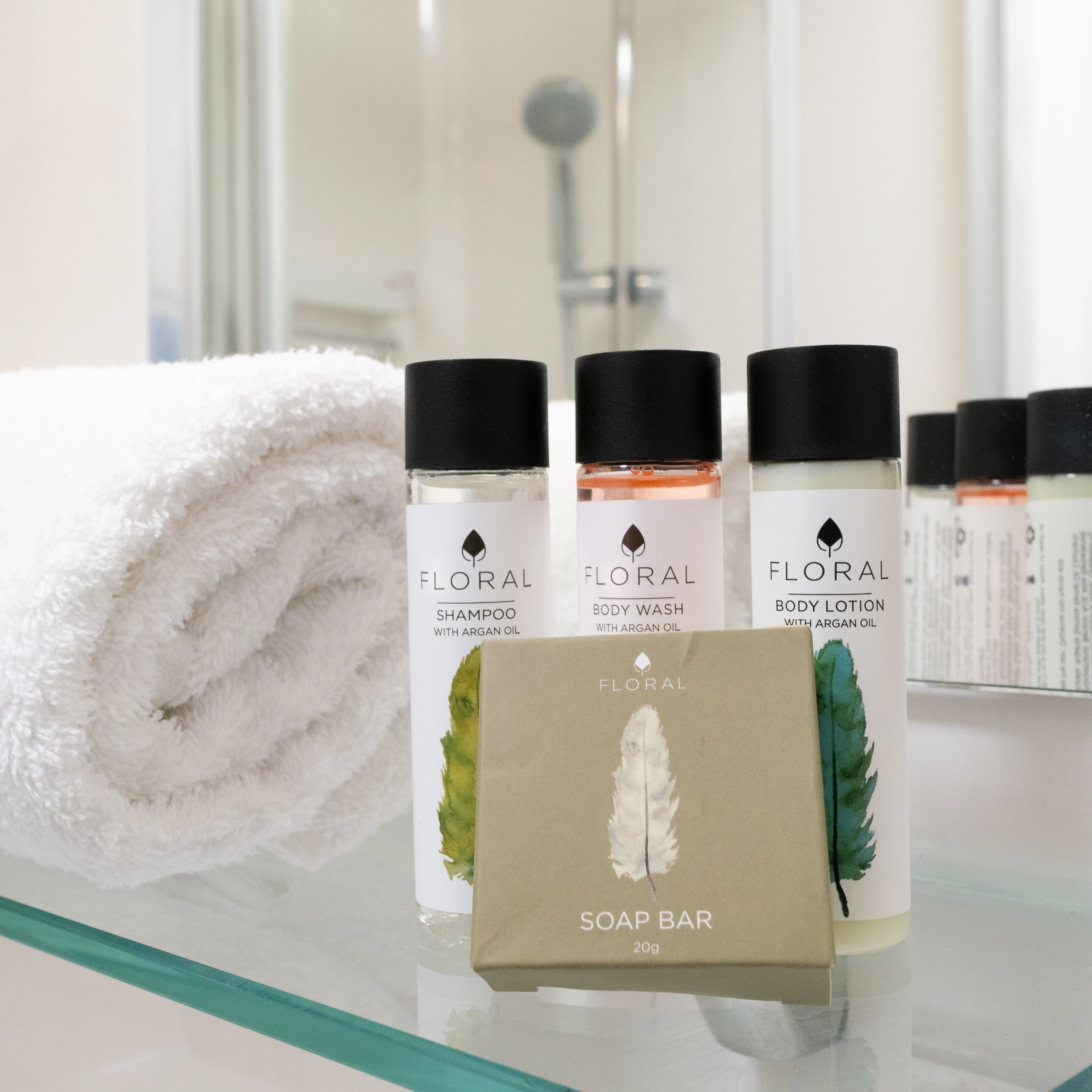 Floral toiletries set with shampoo, body wash, body lotion, and a soap bar on a glass shelf beside a rolled white towel.