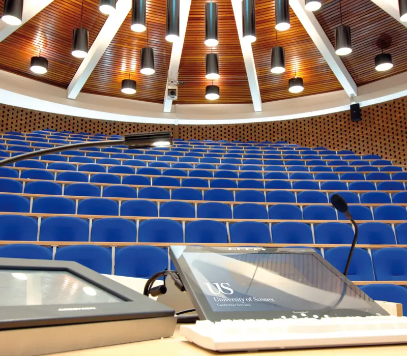 Lecture hall with blue seats, viewed from the podium perspective, featuring a touchscreen labeled "University of Sussex."