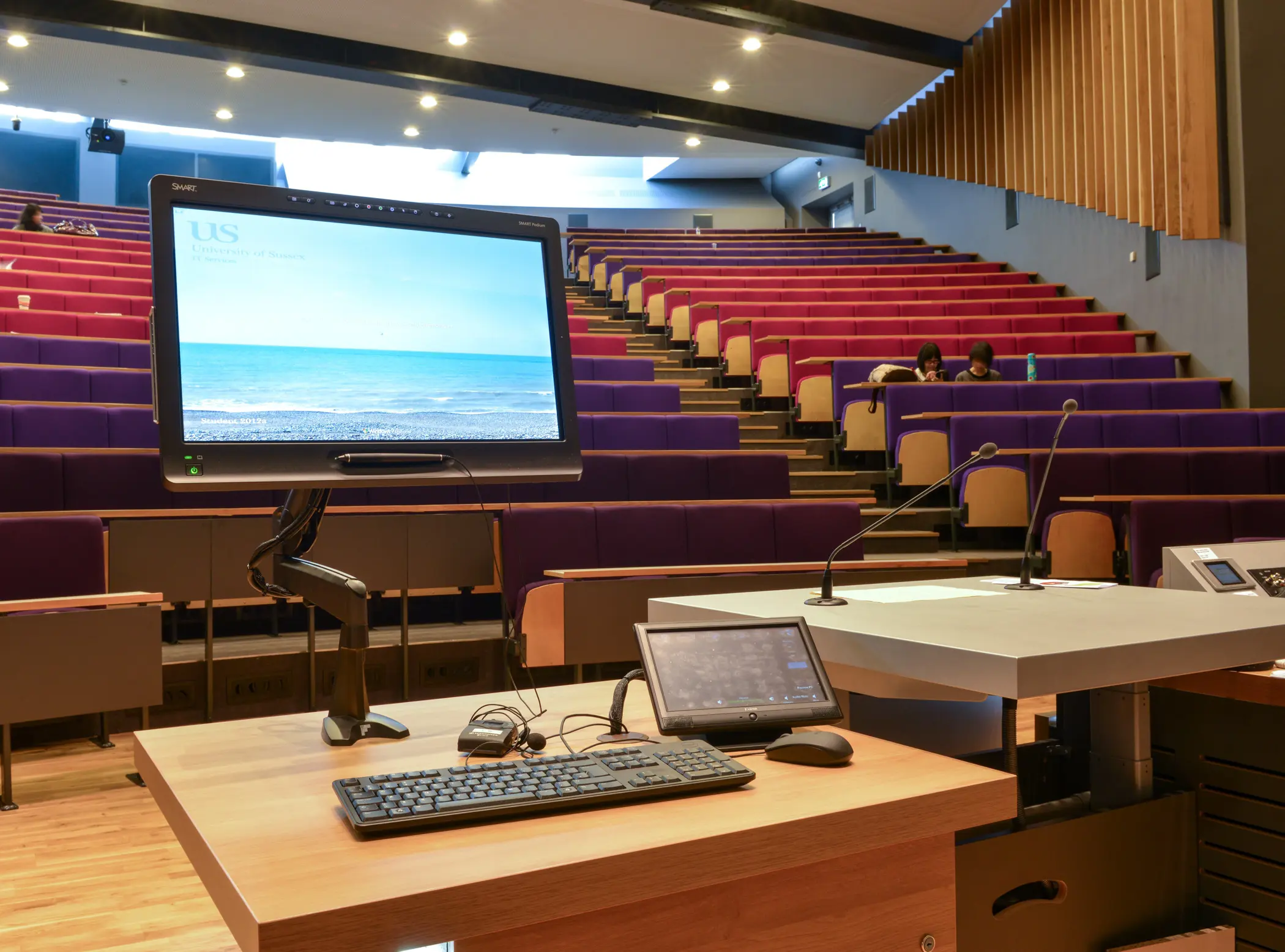 Lecture hall with purple and red seats, view from podium with computer screens and microphones, two people seated.