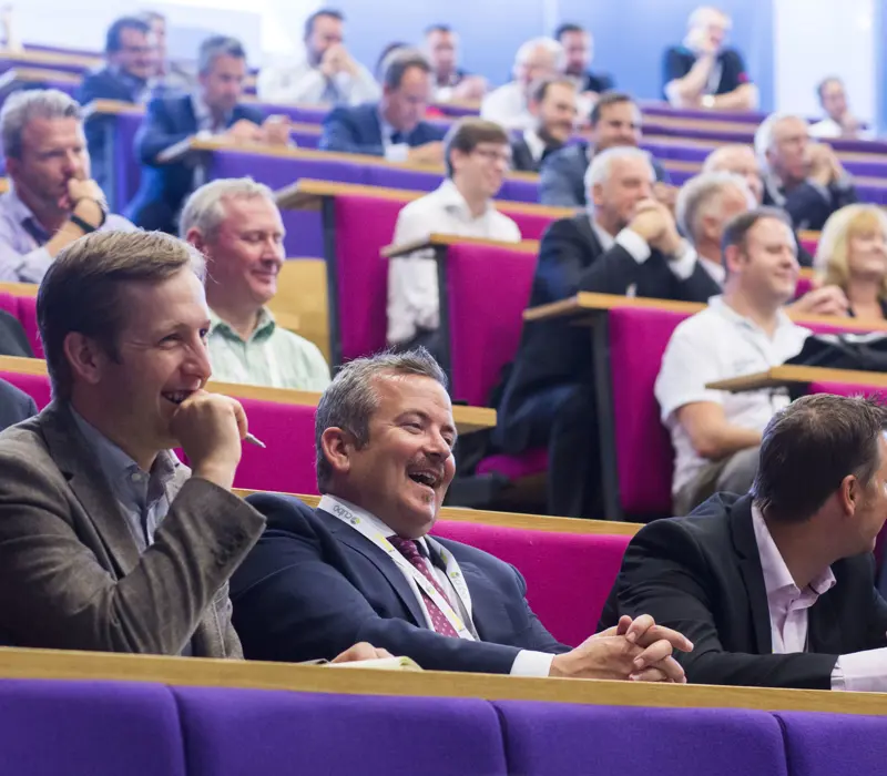 Audience in a lecture hall, smiling and laughing, with rows of people seated in colorful auditorium seats.