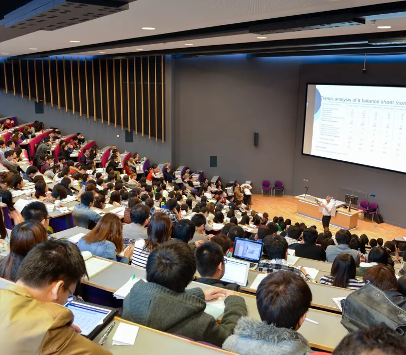 A lecture hall filled with students, some using laptops. A lecturer presents data on a large screen.