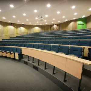 Empty lecture hall with tiered seating, blue chairs, wooden desks, and a green exit door in the background.