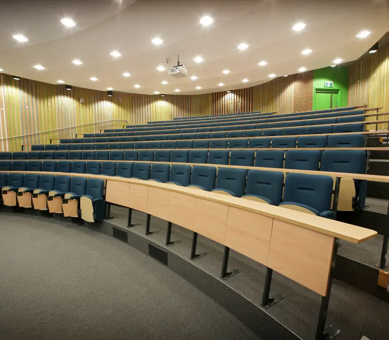 Empty lecture hall with tiered seating, blue chairs, wooden desks, and a green exit door in the background.