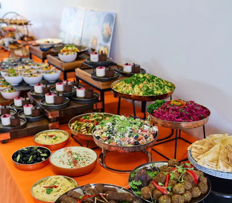 A colorful buffet with various dishes including salads, hummus, olives, falafel, and pita bread on an orange tablecloth.