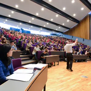 Lecture hall with a professor speaking to a large, engaged audience of students seated in tiered rows.