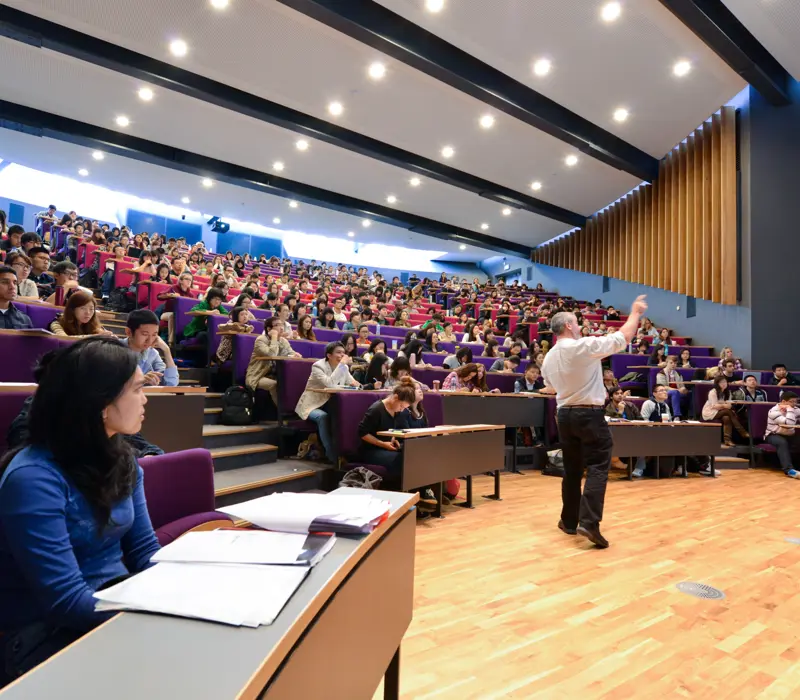 Lecture hall with a professor speaking to a large, engaged audience of students seated in tiered rows.