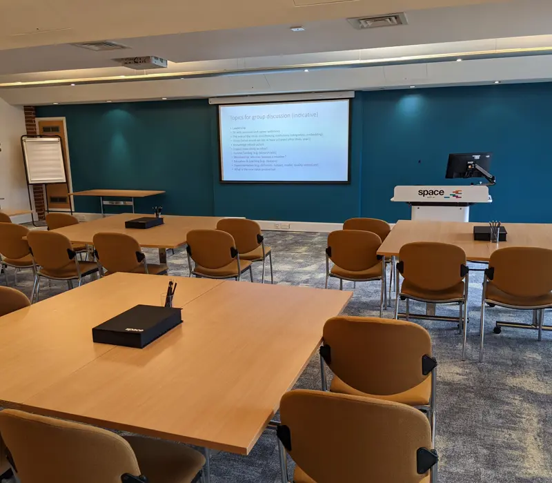 A conference room with arranged chairs, tables, a presentation on a screen, and a podium with a monitor.