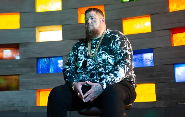 Rag n Bone Man sits in front of a wall with colorful, rectangular glass panels. They wear a patterned jacket and black pants.