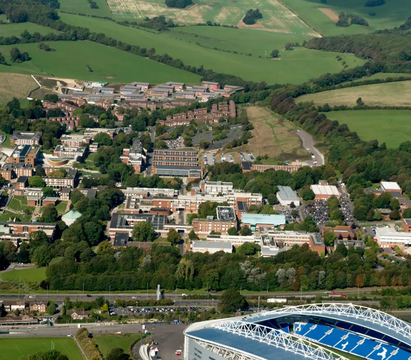 Aerial view of a university campus with multiple buildings surrounded by green fields and trees, adjacent to part of a stadium.