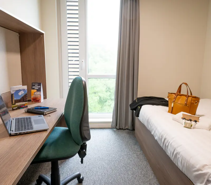 Modern dorm room with a desk, laptop, and chair beside a bed with a handbag and jacket. Large window in the background.
