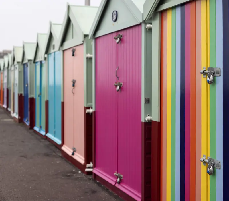 Colorful beach huts in a row, each with a distinct vibrant paint, featuring a rainbow-striped door on the right.