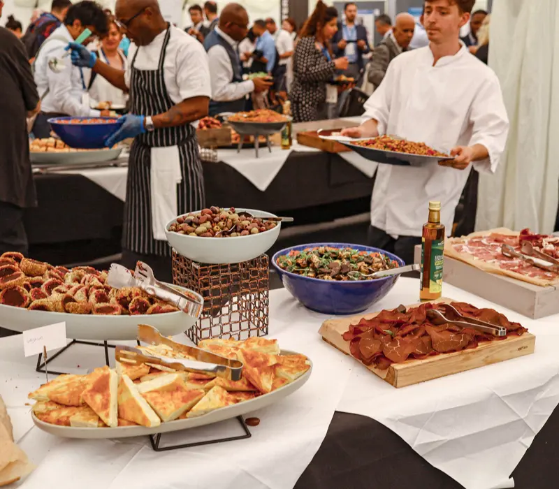 Chefs and a variety of dishes at a bustling buffet; a chef holds a tray of food.