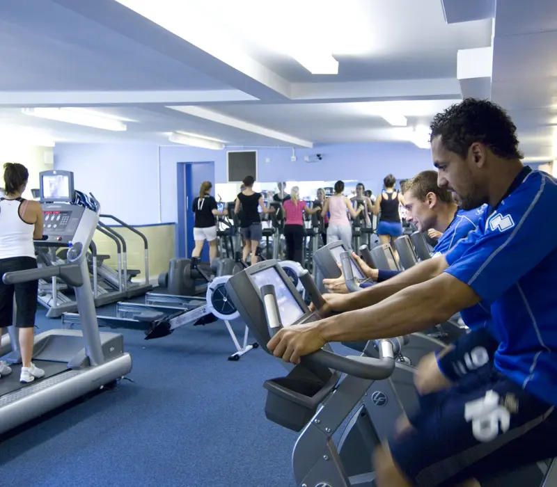 People exercising on stationary bikes and treadmills in a gym with blue carpet and walls.