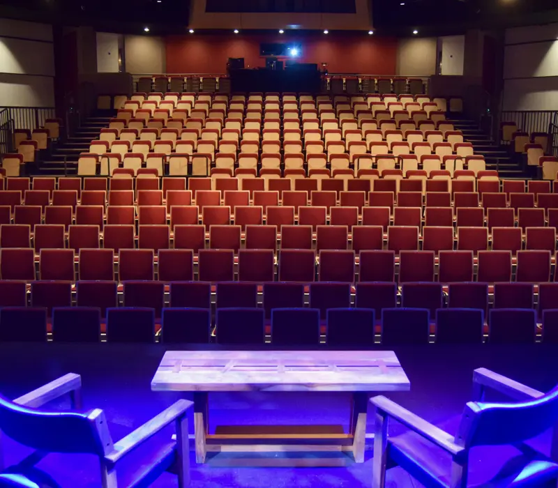Empty theater with rows of red seats, viewed from the stage with two chairs and a table, illuminated by blue lighting.