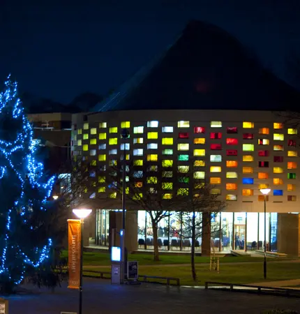 A circular building with colorful lit windows at night next to a tree decorated with blue lights.