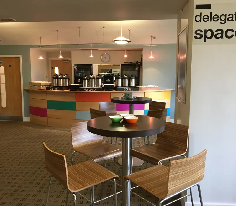 Modern coffee lounge with wooden chairs and tables, multi-colored counter, and coffee dispensers in a welcoming setting.