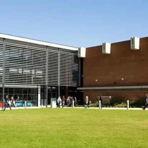 Students walking across a lawn in front of a modern building with glass and brick facade under a clear blue sky.