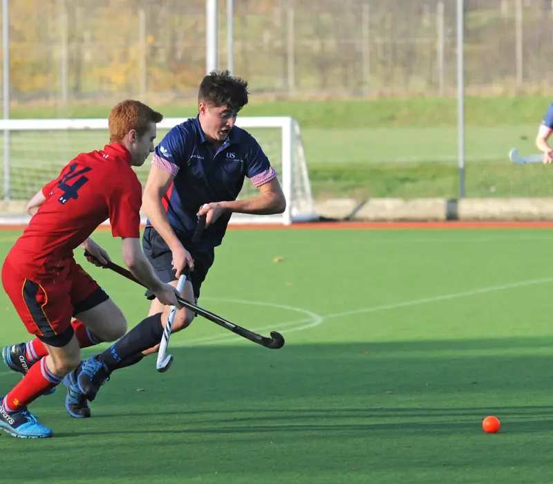 Two field hockey players in action on a green pitch, one in red and the other in blue, chasing an orange ball.