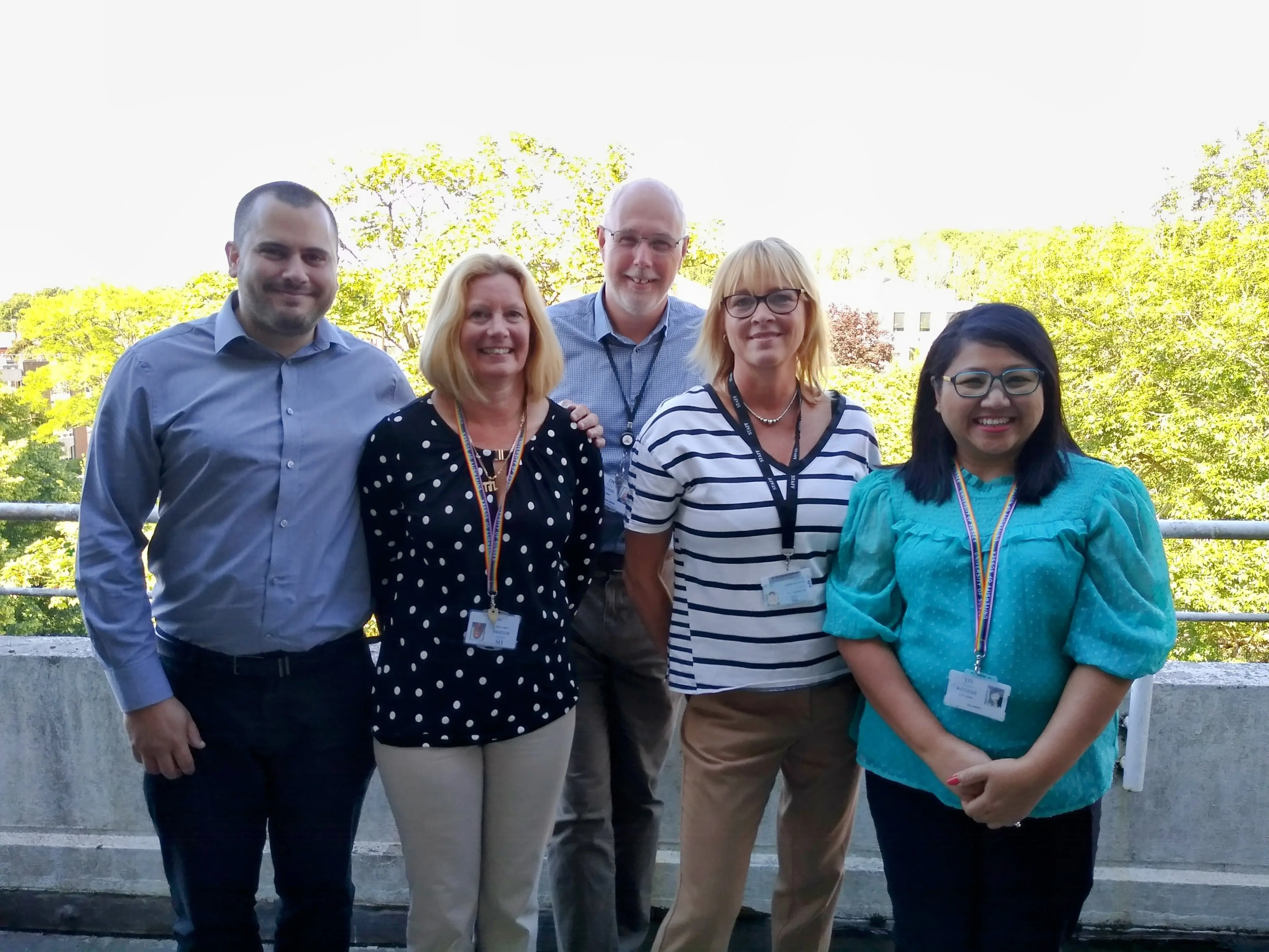 Five people standing outdoors, smiling, wearing ID badges. Trees and a building are visible in the background.