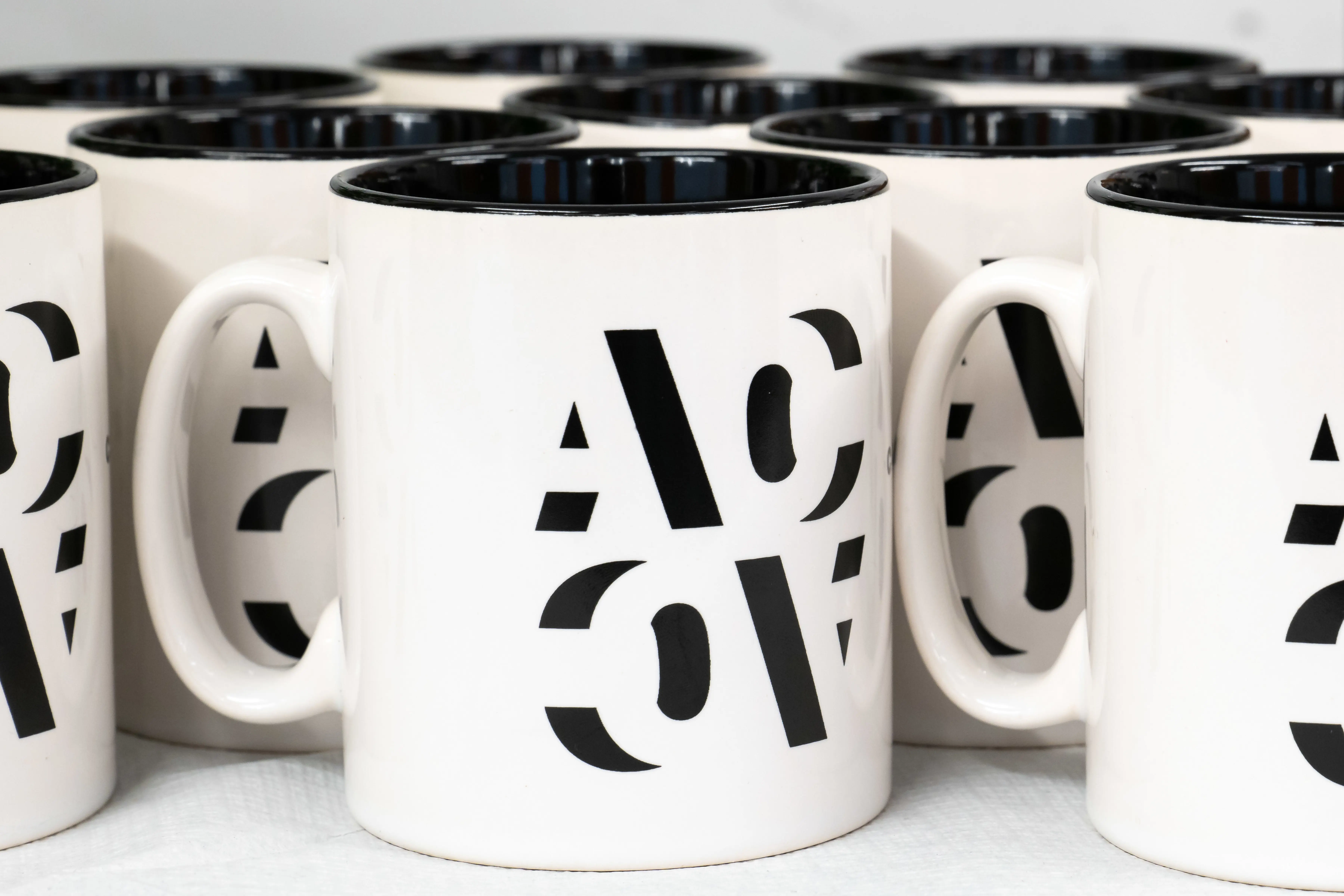 Mugs with bold black "ACO" lettering, neatly arranged in rows.