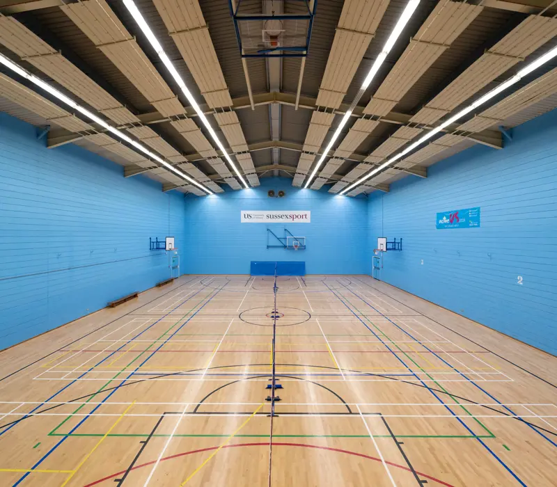 Indoor sports hall with a wooden floor and blue walls, lined for various sports. Basketball hoop and court markers are visible.