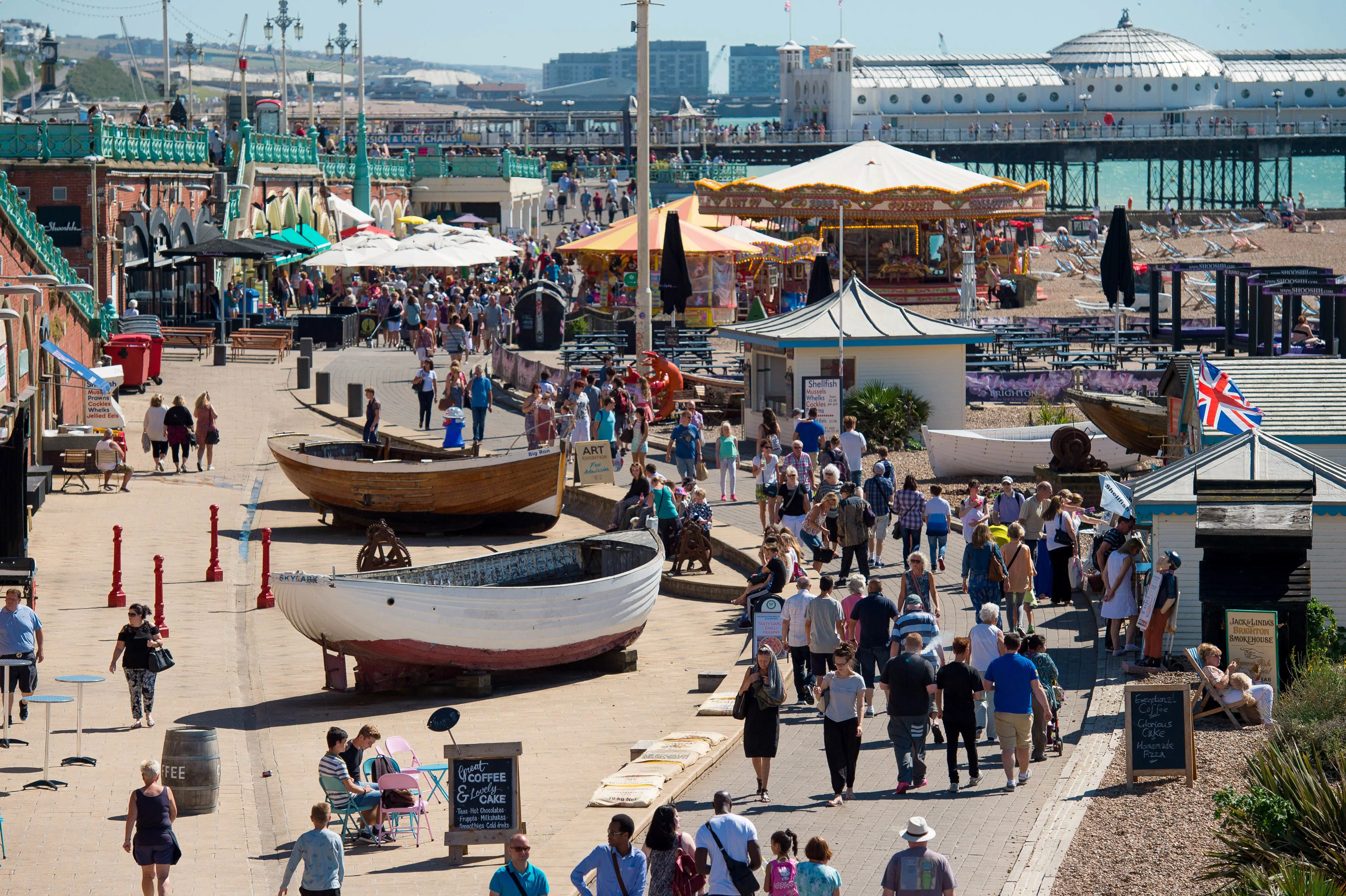 Busy boardwalk by the sea with people, boats, and a carousel; beach and pier in the background on a sunny day.