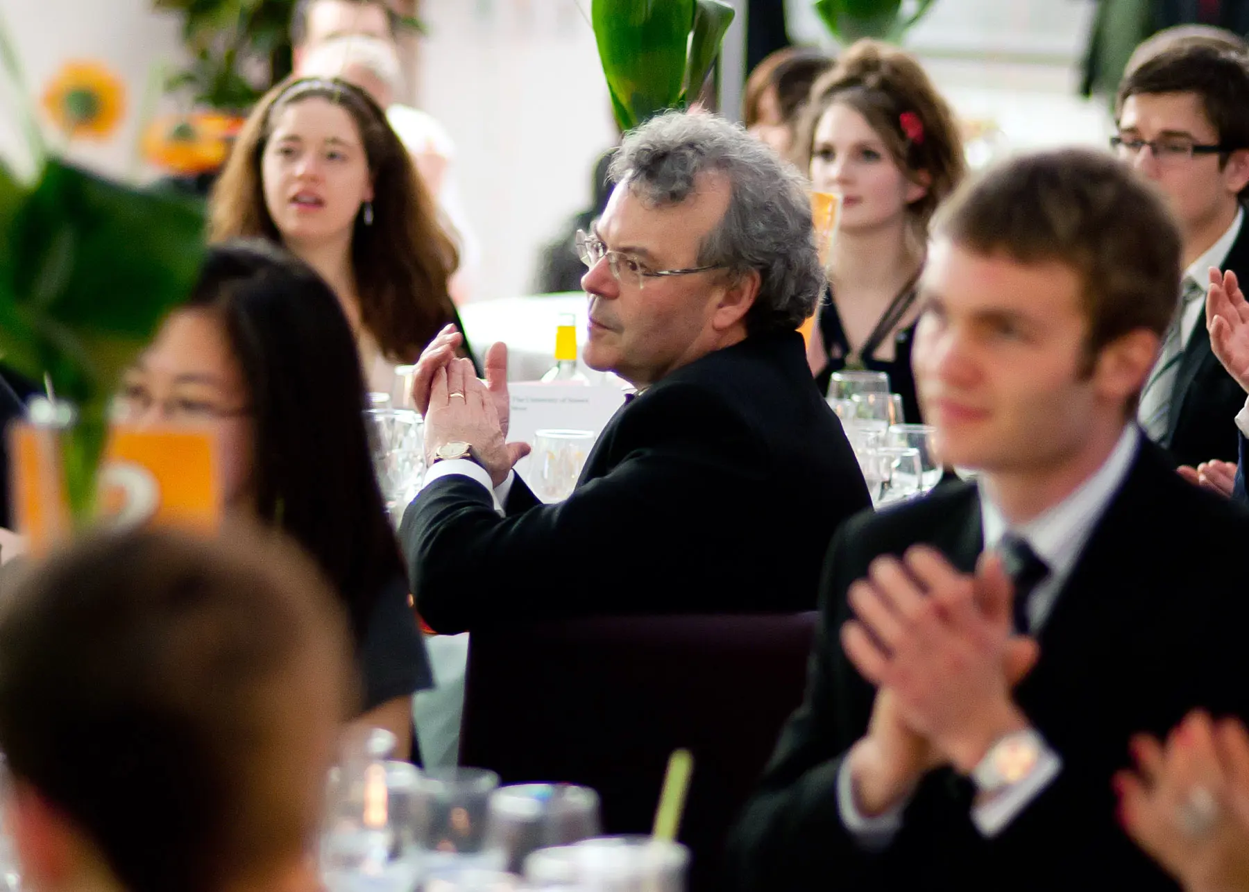 A group of people at a formal event clapping, with a man in a suit in focus, surrounded by glasses and decor.