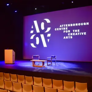 Stage setup with a podium, two chairs, and a table at the Attenborough Centre for the Creative Arts, with a projected logo.