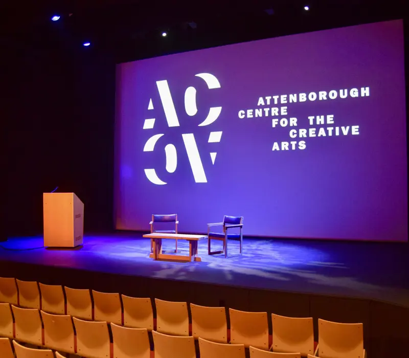 Stage setup with a podium, two chairs, and a table at the Attenborough Centre for the Creative Arts, with a projected logo.