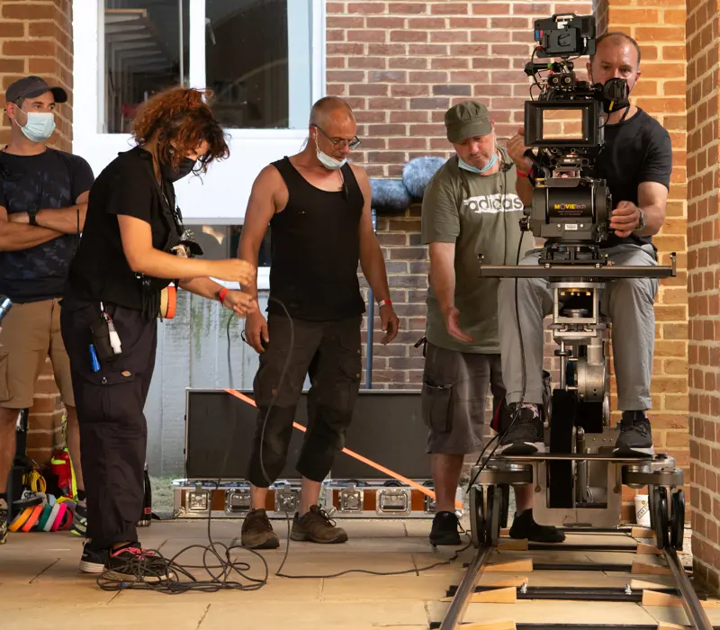 Film crew operating a camera on a dolly track in an outdoor brick corridor, with crew members adjusting equipment.
