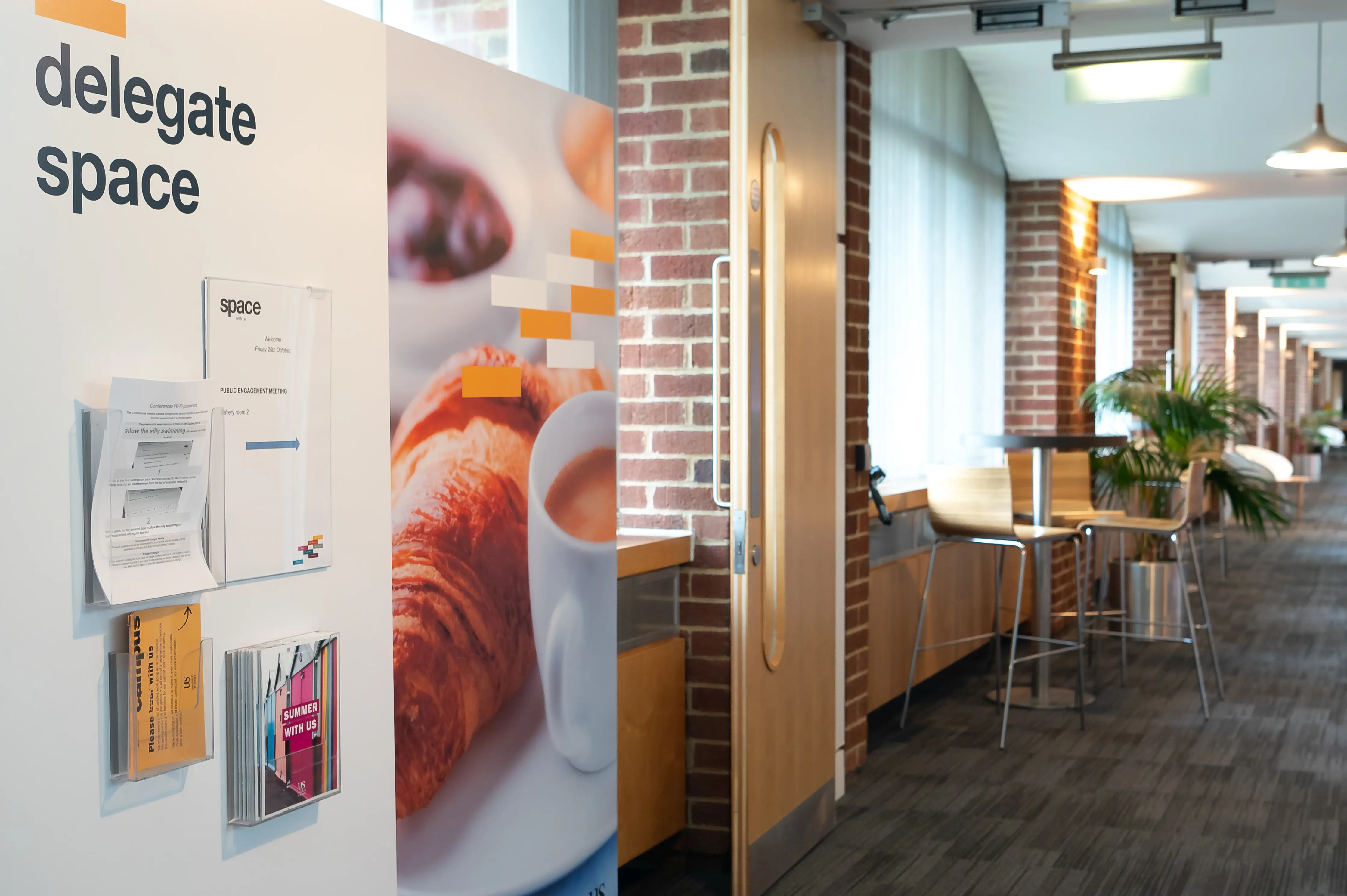 Modern office hallway with a coffee and croissants poster, brick walls, and wooden chairs near a window.