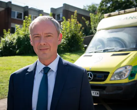 John Simm in a blue suit and teal tie stands outdoors in front of a yellow ambulance, with a building and greenery in the background.