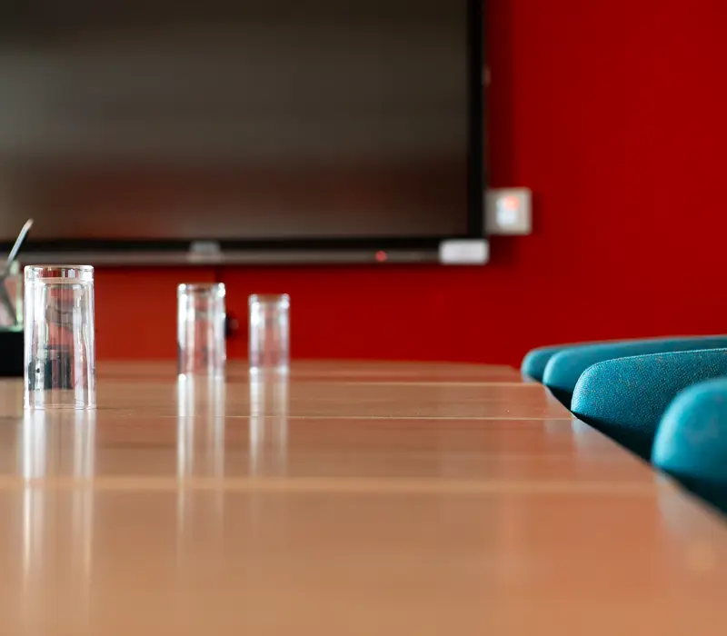 Empty conference room with blue chairs, wooden table, three glasses, and a large screen on a red wall.