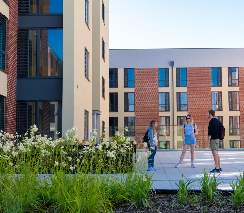 Three people converse in a courtyard surrounded by modern buildings and greenery on a sunny day.