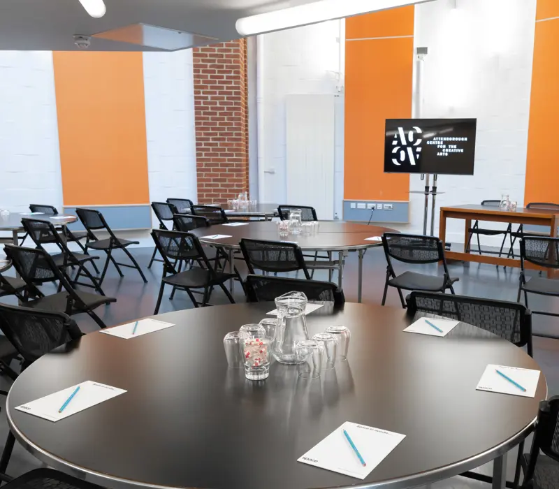 Modern conference room with round tables, chairs, notebooks, and pens. A screen and notepads are set up for a meeting.