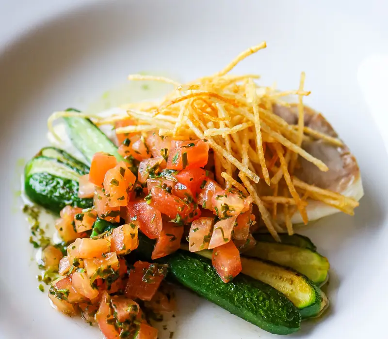 Gourmet dish with sliced zucchini, diced tomatoes, herbs, and crispy fries on a white plate.
