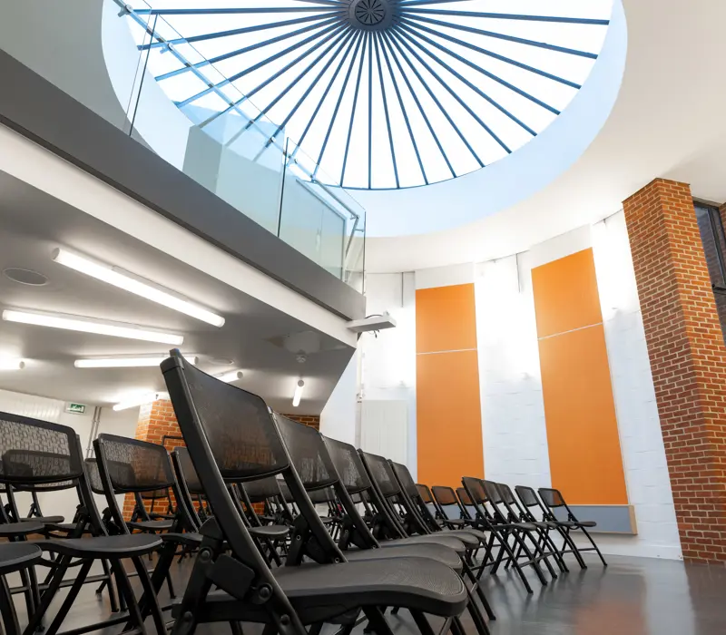 Modern auditorium with gray folding chairs, white walls, orange panels, and a circular skylight with a radial pattern.
