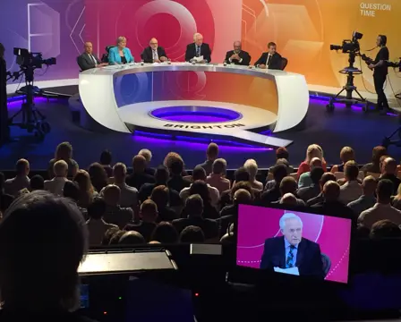 A Question Time TV panel discussion takes place on stage with six people seated at a curved table, facing a live audience in a studio.