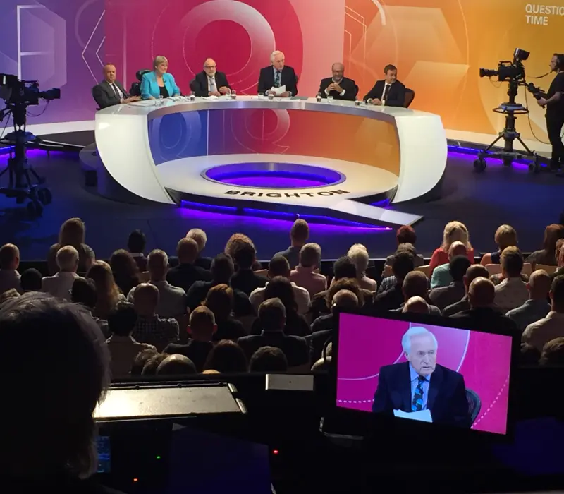 A Question Time TV panel discussion takes place on stage with six people seated at a curved table, facing a live audience in a studio.